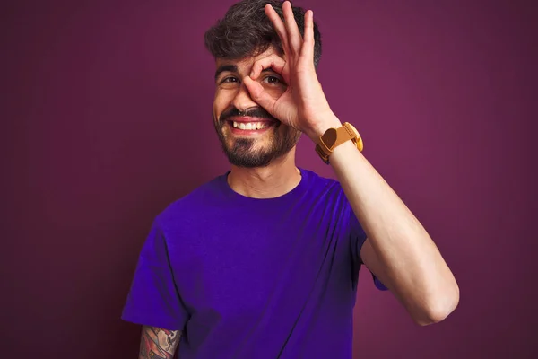 孤立した紫色の背景の上に立ってタトゥーを着た若い男は 手の笑顔でOkジェスチャーを行い 幸せな顔で指を通して目を見ます — ストック写真