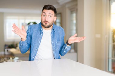 Genç yakışıklı adam evde clueless ve şaşkın bir ifadeyle kollar ve eller kaldırdı. Şüphe kavramı.
