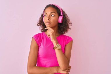 Genç Brezilyalı kadın izole pembe arka plan üzerinde kulaklık kullanarak müzik dinlemek soru hakkında düşünme ciddi yüz, çok karışık fikir