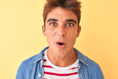 İzole sarı arka plan üzerinde çizgili t-shirt ve kot gömlek giyen genç yakışıklı adam sürpriz bir yüz ile şok korkmuş, korku ifade korku ve heyecanlı