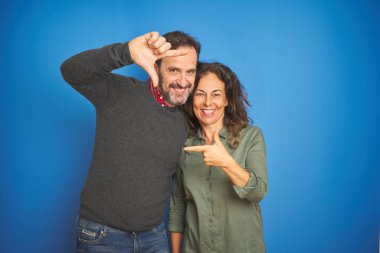 Güzel orta yaş çift birlikte izole mavi arka plan gülümseyen el ve parmaklarla mutlu yüz ile çerçeve yapma üzerinde ayakta. Yaratıcılık ve Fotoğrafçılık konsepti.