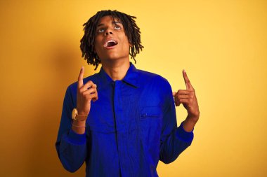 İzole sarı arka plan üzerinde mekanik üniforma giyen dreadlocks ile Afro işçi adam hayret ve görünümlü ve parmaklar ve yükseltilmiş kollar ile işaret şaşırttı.
