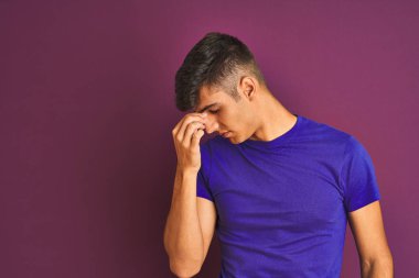 İzole mor arka plan üzerinde duran t-shirt giyen genç hintli adam yorgun burun ve gözleri sürtünme yorgunluk ve baş ağrısı hissi. Stres ve hayal kırıklığı kavramı.