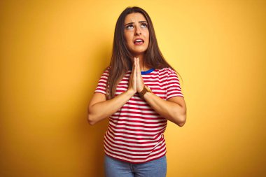 İzole sarı arka plan üzerinde ayakta çizgili t-shirt giyen genç güzel kadın yalvarıyor ve çok duygusal ve endişeli yüzünde umut ifade ile birlikte eller ile dua. Affedilmesini istiyorum. Din konsepti.