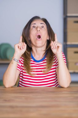 Orta yaş kıdemli kadın evde masada oturan şaşırdı ve bakmak ve parmaklar ve yükseltilmiş silah ile işaret şaşırttı.