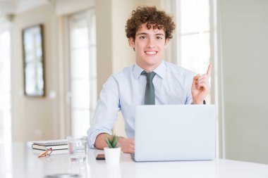 Yüz üzerinde büyük bir gülümseme ile ofiste bilgisayar laptop ile çalışan genç iş adamı, kamera bakarak yan el ve parmak ile işaret.