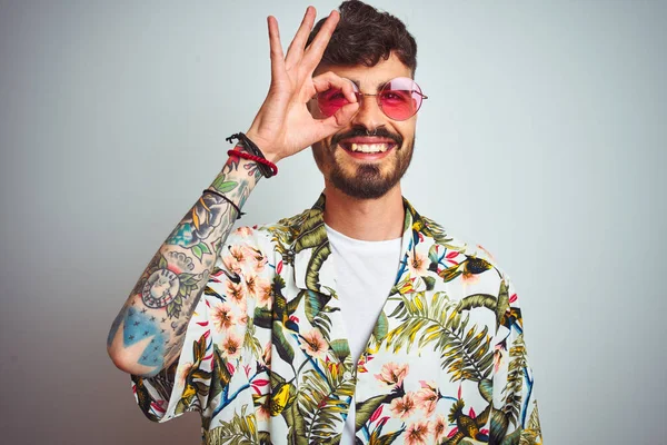 Man with tattoo on vacation wearing summer shirt sunglasses over isolated white background doing ok gesture with hand smiling, eye looking through fingers with happy face.
