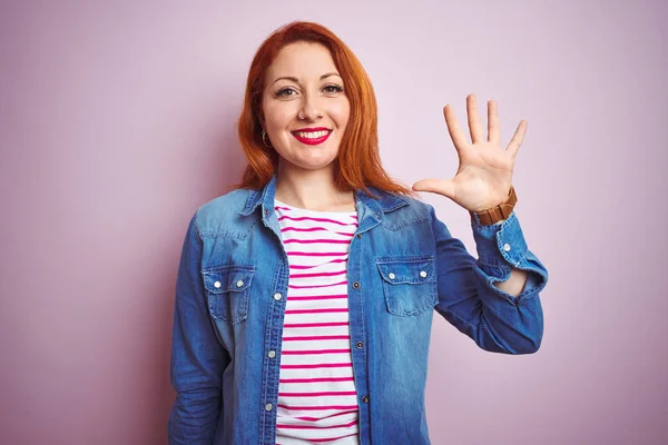 デニムシャツとストライプのTシャツを着た美しい赤毛の女性は 自信を持って幸せに微笑みながら 指番号5を示し 指で指を上げる孤立したピンクの背景の上に — ストック写真