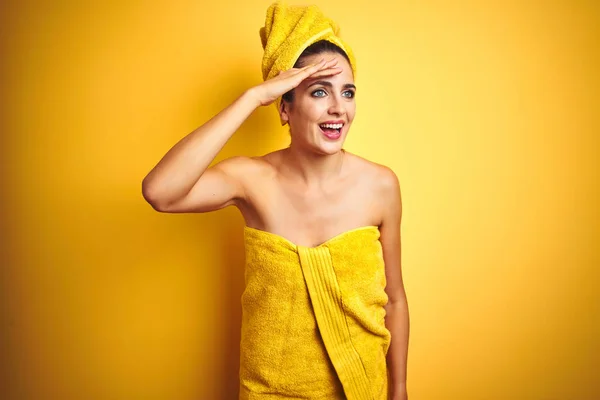 Beautiful woman wearing shower towel on body and head over yellow isolated background very happy and smiling looking far away with hand over head. Searching concept.