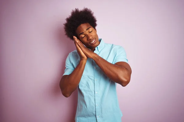 Young american man with afro hair wearing blue shirt standing over isolated pink background sleeping tired dreaming and posing with hands together while smiling with closed eyes.