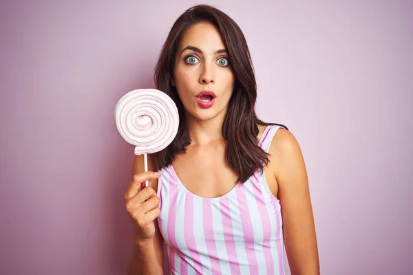 ピンクの孤立した背景の上に甘いキャンディを食べる若い美しい女性は 驚きの顔でショックを受け 恐怖の表情で興奮 — ストック写真