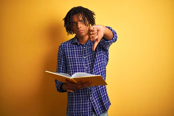 Afro american student man with dreadlocks reading book over isolated yellow background with angry face, negative sign showing dislike with thumbs down, rejection concept