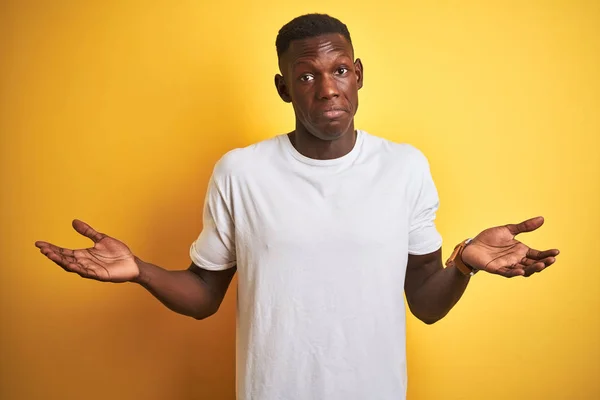 孤立した黄色の背景の上に立つ白いTシャツを着た若いアフリカ系アメリカ人男性は 腕と手を上げて混乱した表情を浮かべた 疑いの概念 — ストック写真
