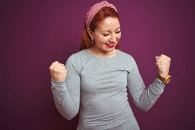 Gri t-shirt ve diadem üzerinde izole mor arka plan giyen güzel kızıl saçlı kadın çok mutlu ve heyecanlı silah ile kazanan jest yaparak, gülümseyen ve başarı için çığlık kaldırdı. Kutlama konsepti.