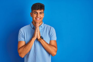 Yalnız mavi arka plan üzerinde duran rahat polo giyen genç hintli adam elleri birlikte kendinden emin gülümseyerek af dileyen dua.