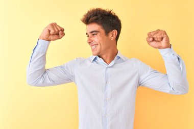 Genç yakışıklı işadamı izole sarı arka plan üzerinde zarif gömlek giyen kol kasları gururla gülümseyerek gösteren. Fitness konsepti.