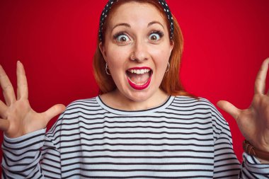 İzole kırmızı arka plan üzerinde ayakta çizgili lacivert tişört giyen güzel Redhead kadın deli kutluyor ve başarı için hayran silah kaldırdı ve açık gözleri heyecanlı çığlık. Kazanan kavramı