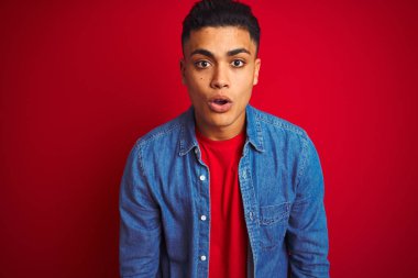 İzole kırmızı arka plan üzerinde ayakta kot gömlek giyen genç Brezilyalı adam korkuyor ve sürpriz ifade, korku ve heyecanlı yüz ile şok.
