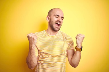 Sarı izole arka plan üzerinde rahat çizgili t-shirt giyen sakal ile genç kel adam şaşırttı kutluyor ve başarı için hayran kollarını kaldırdı ve gözleri kapalı. Kazanan kavramı.
