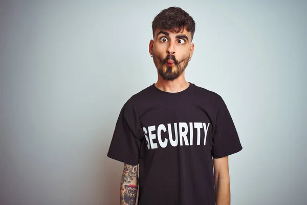 Young safeguard man with tattoo wering security uniform over isolated white background making fish face with lips, crazy and comical gesture. Funny expression.
