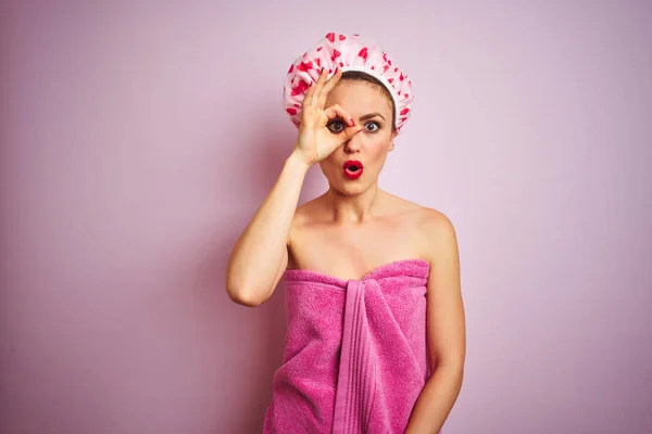 年轻美丽的女人穿着毛巾和浴帽淋浴后 在粉红色孤立的背景做Ok手势震惊惊讶的脸 眼睛透过手指看 不信的表达 — 图库照片