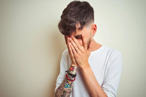 Young man with tattoo wearing t-shirt standing over isolated white background with sad expression covering face with hands while crying. Depression concept.