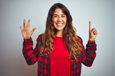 Beyaz izole arka plan üzerinde ayakta kırmızı t-shirt ve ceket giyen genç güzel kadın gösteren ve parmak numarası altı ile işaret kadar emin ve mutlu gülümseyen.