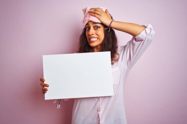Genç Kadın Pijama ve izole pembe arka plan üzerinde uyku maskesi tutan afiş giyen el baş ile, utanç ve sürpriz yüz, öfkeli ve sinirli ile şok vurguladı. Korku ve hata için üzgün.