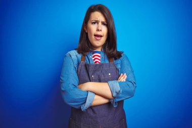 Mavi izole arka plan üzerinde mağaza üniforma apron giyen genç güzel iş kadın şüpheci ve sinir, Çapraz kollar ile yüz üzerinde ifade onaylamıyor. Negatif kişi.