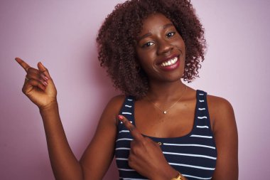 Genç Afrikalı Afro kadın izole pembe arka plan üzerinde ayakta çizgili t-shirt giymiş ve iki el ve parmak tarafına işaret kamera bakıyor.