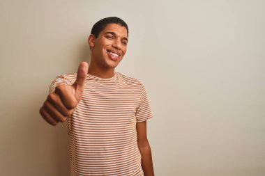 Genç yakışıklı Arap adam izole beyaz arka plan üzerinde çizgili t-shirt giyen el ile jest kadar mutlu başparmak yapıyor. Başarı göstererek kameraya bakan ifadeyi onaylama.