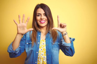 Sarı izole arka plan üzerinde ayakta genç güzel kadın gösteren ve parmaklarının sayısı yedi ile işaret kadar güven ve mutlu gülümseyen.
