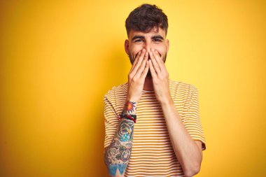 Dövme ile genç adam izole sarı arka plan üzerinde duran çizgili t-shirt ayakta gülüyor ve eller, dedikodu ve skandal kavramı ile ağız kapsayan utanmış kıkırdayan