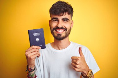 Genç adam dövme ile izole sarı arka plan üzerinde Alman Almanya pasaport giyen büyük gülümseme ok işareti yapıyor, parmaklar ile başparmak, mükemmel işareti