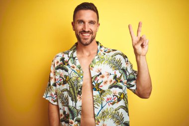 Sarı izole arka plan üzerine yaz Hawaii çiçekler gömlek giyen yakışıklı adam mutlu yüz zafer işareti yapıyor kamera göz kırpıyor ile gülümsüyor. İki numara.