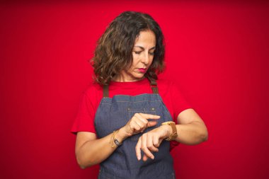 Kırmızı izole arka planda apron üniforma giyen orta yaş kıdemli kadın bilek saatindeki saati kontrol etme, rahat ve emin