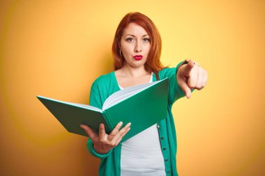 Genç Redhead öğrenci kadın kamera ve size, el işareti, önde pozitif ve emin jest parmak ile işaret sarı izole arka plan üzerinde yeşil kitap okuma