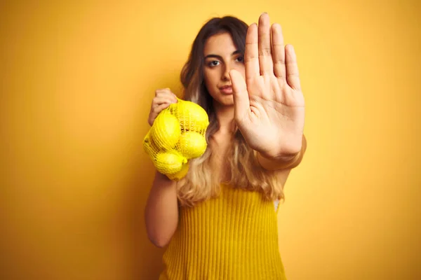 真面目で自信に満ちた表情でストップサインをする開いた手で黄色の孤立した背景の上にレモンのネットを保持する若い美しい女性 防衛ジェスチャー — ストック写真