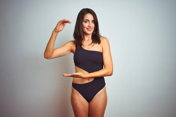 Beautiful woman wearing bikini swimwear over white isolated background gesturing with hands showing big and large size sign, measure symbol. Smiling looking at the camera. Measuring concept.