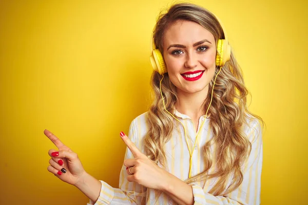黄色の孤立した背景の上にヘッドフォンを使用して音楽を聴く若い美しい女性は微笑み 側面に両手と指で指してカメラを見て — ストック写真