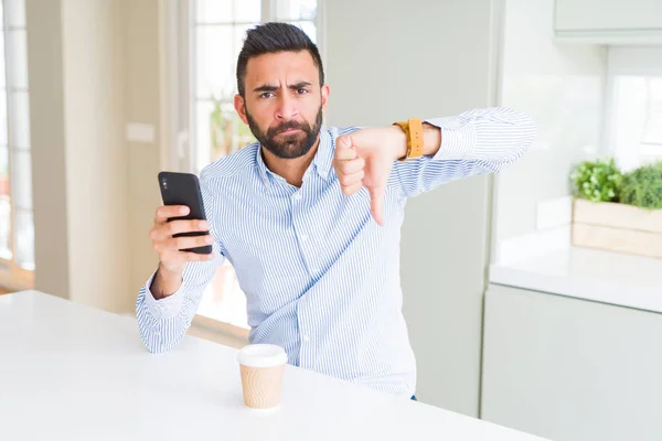 Handsome hispanic business man drinking coffee and using smartphone with angry face, negative sign showing dislike with thumbs down, rejection concept