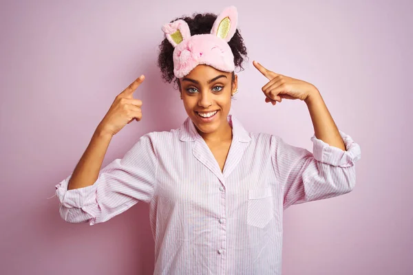 年轻的非洲裔美国妇女穿着睡衣和面具在孤立的粉红色背景微笑指向头部与双手手指 伟大的想法或思想 良好的记忆力 — 图库照片