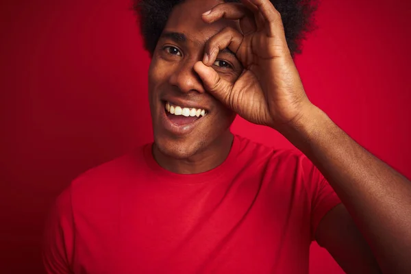 Young american man with afro hair wearing t-shirt standing over isolated red background with happy face smiling doing ok sign with hand on eye looking through fingers