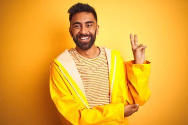 Genç Hint adam kamera zafer işareti yapıyor mutlu yüz göz kırpıyor ile gülümseyen izole sarı arka plan üzerinde ayakta yağmurluk giyiyor. İki numara.