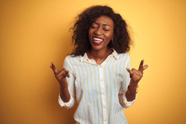 Çizgili gömlek giyen Afro-amerikan kadın izole sarı arka plan üzerinde duran deli ifade elleri ile rock sembolü yapıyor bağırarak. Müzik yıldızı. Ağır konsept.