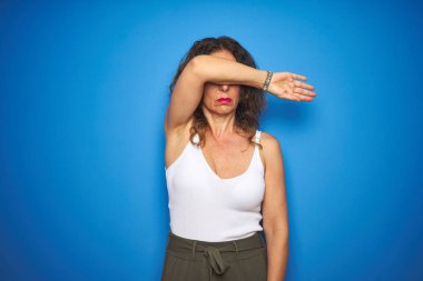 Orta yaş yaşlı kadın kıvırcık saçlı mavi izole arka planda ayakta kol ile gözleri kapsayan, ciddi ve üzücü görünümlü. Görüsüz, gizleme ve reddetme konsepti