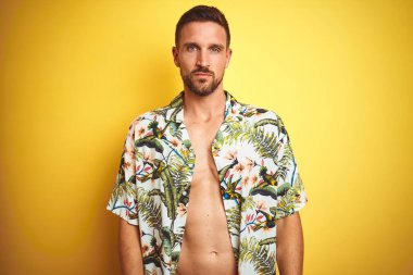 Yüze ciddi ifade ile sarı izole arka plan üzerinde yaz Hawaii çiçekler gömlek giyen yakışıklı adam. Basit ve doğal kamera bakıyor.