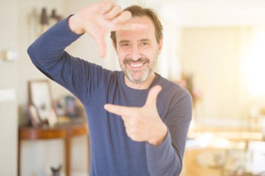Yakışıklı orta yaş erkek evde elleriyle yapım çerçeve ve parmakları ile mutlu yüz gülümsemeye. Yaratıcılık ve Fotoğrafçılık kavramı.
