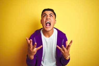 Genç Brezilyalı adam izole sarı arka plan üzerinde ayakta mor sweatshirt giyen çılgın ve deli çığlık ve agresif ifade ve silah ile bağırdı kaldırdı. Hayal kırıklığı kavramı.