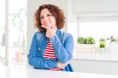 Evde kot ceket giyen kıvırcık saçlı orta yaş kıdemli kadın çapraz silah ve el çene üzerinde yükseltilmiş ile gülümseyen kamera emin görünümlü. Pozitif düşünme.